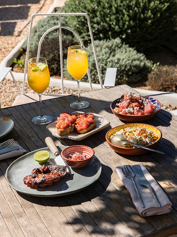 The image features an inviting spread of dishes on an outdoor table at a restaurant in Menorca. The setting includes two glasses of vibrant orange cocktails garnished with lime, alongside plates of various tapas like grilled meat with a lime wedge, tomato-topped bread, a fresh watermelon salad with crumbled cheese, and a colorful quinoa salad. The wooden table and surrounding greenery create a relaxed, sunlit dining experience, perfect for enjoying Mediterranean flavors al fresco.