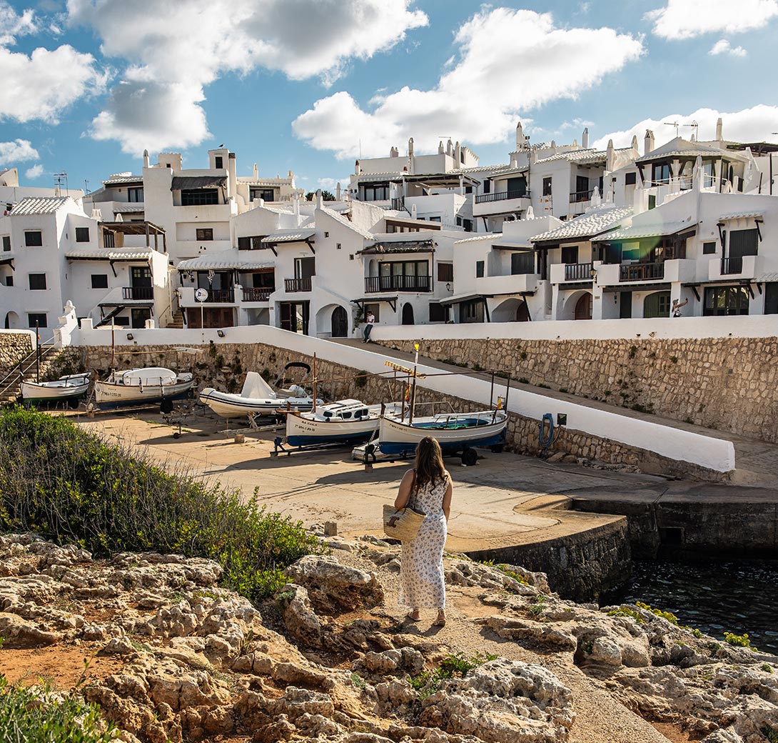 The image captures a charming scene of Binibeca Vell, a traditional whitewashed fishing village in Menorca. The narrow cobblestone streets are lined with picturesque white houses, featuring wooden balconies and quaint architectural details. In the foreground, a woman in a light summer dress walks towards a small marina area where boats are docked, with the rocky shore and clear blue sky enhancing the idyllic Mediterranean atmosphere.