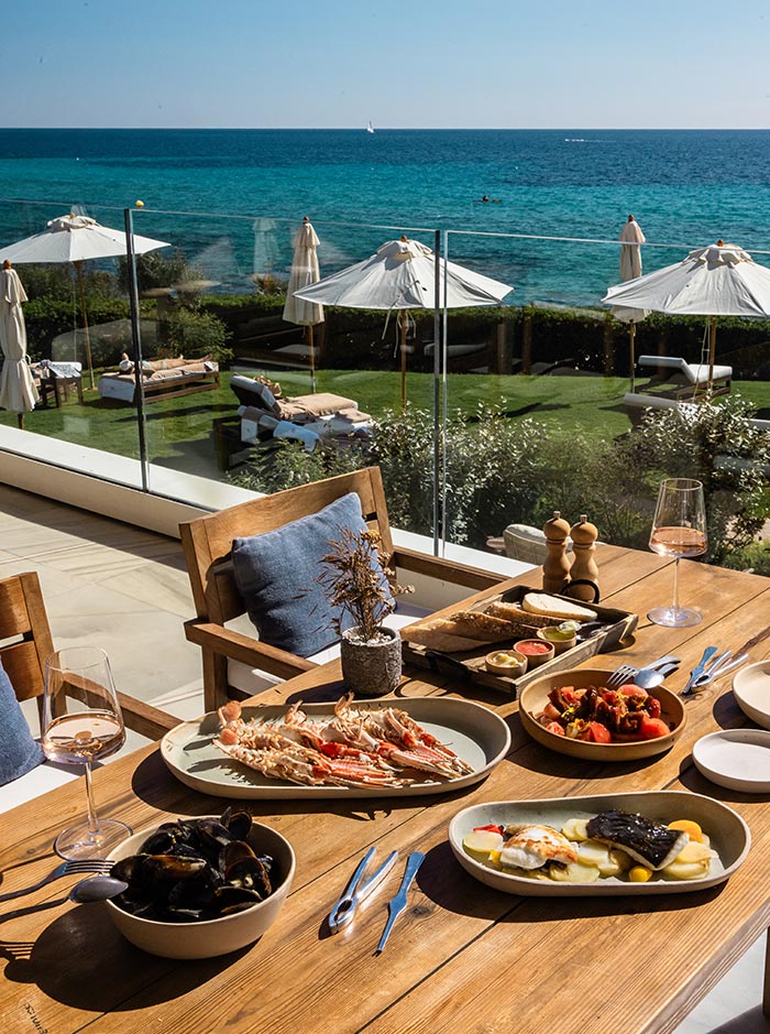 The image showcases a luxurious dining setup at Villa Le Blanc, with a stunning view of the turquoise Mediterranean Sea in the background. The table is adorned with various seafood dishes, including a plate of grilled shellfish, a bowl of mussels, and a beautifully plated fish dish. Accompanying the seafood are fresh bread, dips, and a vibrant tomato salad. The scene is bathed in sunlight, with white parasols and plush lounge chairs visible on the lawn below, creating an elegant and relaxing coastal dining experience.