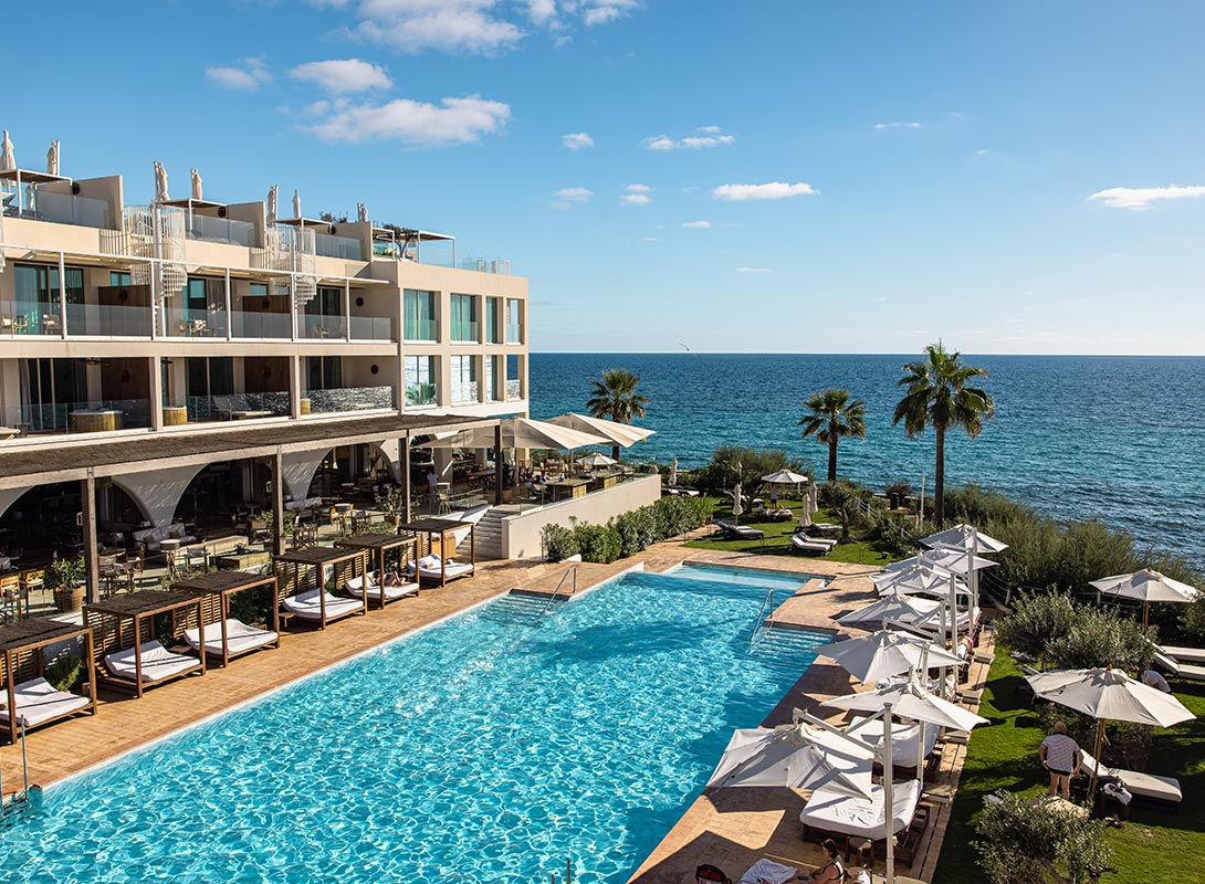 The image captures a luxurious poolside scene at Villa Le Blanc in Menorca, with the bright blue Mediterranean Sea as a backdrop. The modern hotel features a large, sparkling pool surrounded by shaded cabanas and sun loungers, offering guests a perfect spot to relax. Elegant outdoor dining areas and terraces overlook the sea, while palm trees and manicured greenery enhance the resort's serene, coastal atmosphere. The scene is bathed in warm sunlight, creating an inviting and tranquil setting.