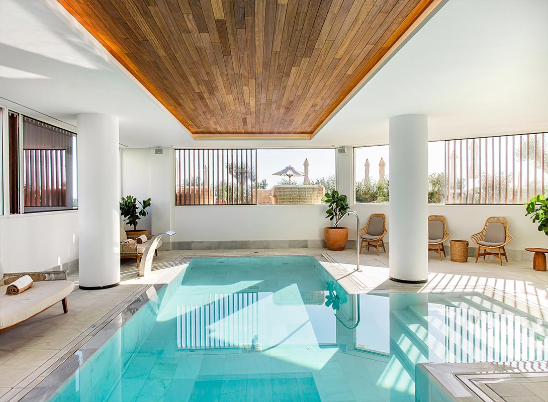 The image shows an elegant indoor spa area at Villa Le Blanc in Menorca, featuring a serene swimming pool surrounded by modern decor. The ceiling has warm wood paneling that contrasts with the white walls, creating a cozy yet airy ambiance. Natural light filters in through large windows, offering a glimpse of outdoor greenery and sun umbrellas. The space is adorned with potted plants and comfortable seating, providing a tranquil setting for relaxation and rejuvenation.