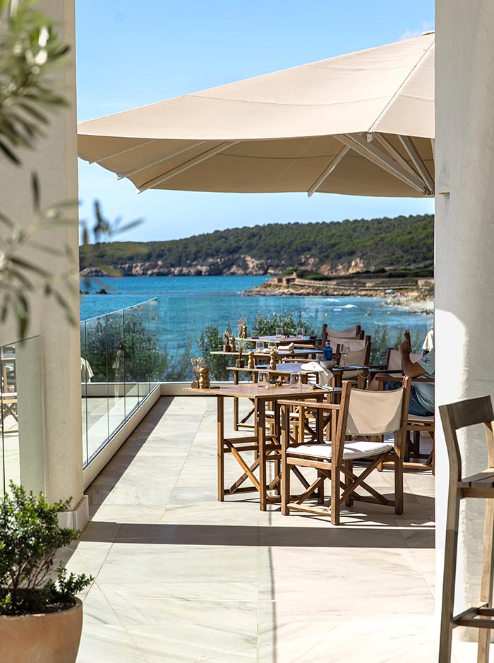 The image depicts an elegant outdoor dining area at Villa Le Blanc, Menorca, overlooking a stunning coastal view. Wooden chairs with light cushions are arranged around tables under large beige sun umbrellas, offering a comfortable and stylish space to enjoy a meal. The scene is framed by glass railings, allowing unobstructed views of the turquoise sea and the rocky shoreline in the distance. The bright, sunny day enhances the relaxed and sophisticated atmosphere of this seaside retreat.