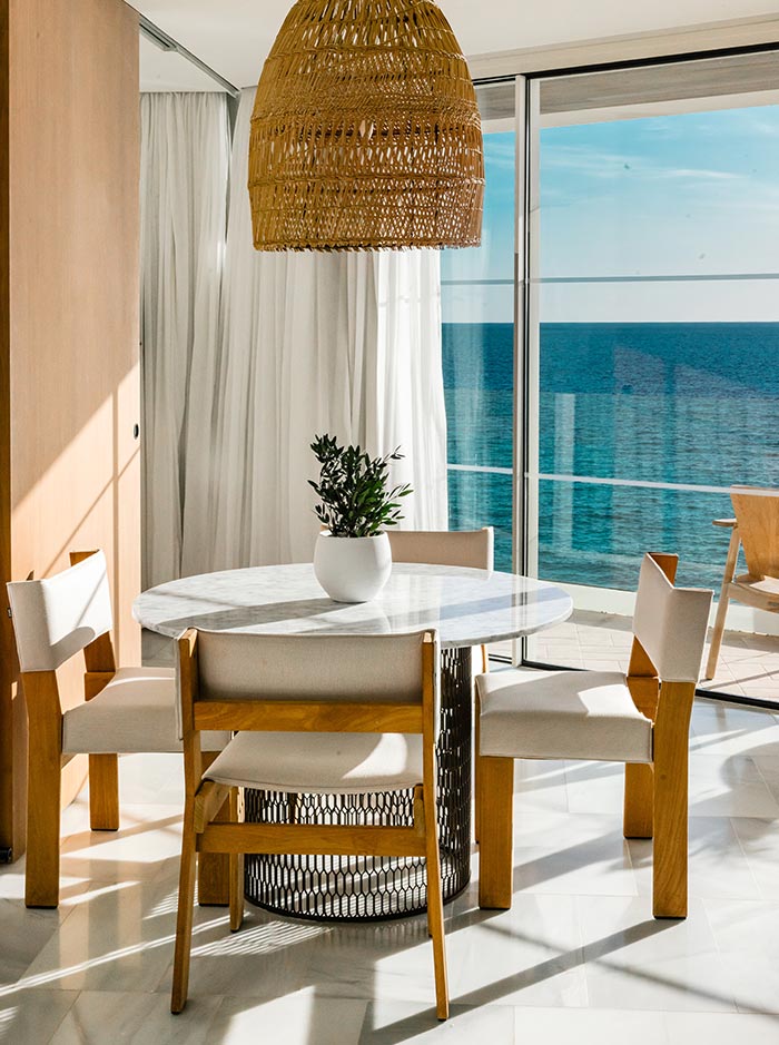 The image showcases a stylish interior of a suite at Villa Le Blanc, Menorca, with a modern, minimalistic design. A round marble table with light wooden chairs is placed near floor-to-ceiling glass doors that open up to a stunning ocean view. Above the table hangs a woven rattan pendant light, adding a natural touch to the space. The bright sunlight fills the room, highlighting the clean lines, soft textures, and the inviting coastal ambiance. A potted plant on the table adds a touch of greenery to the serene setting.