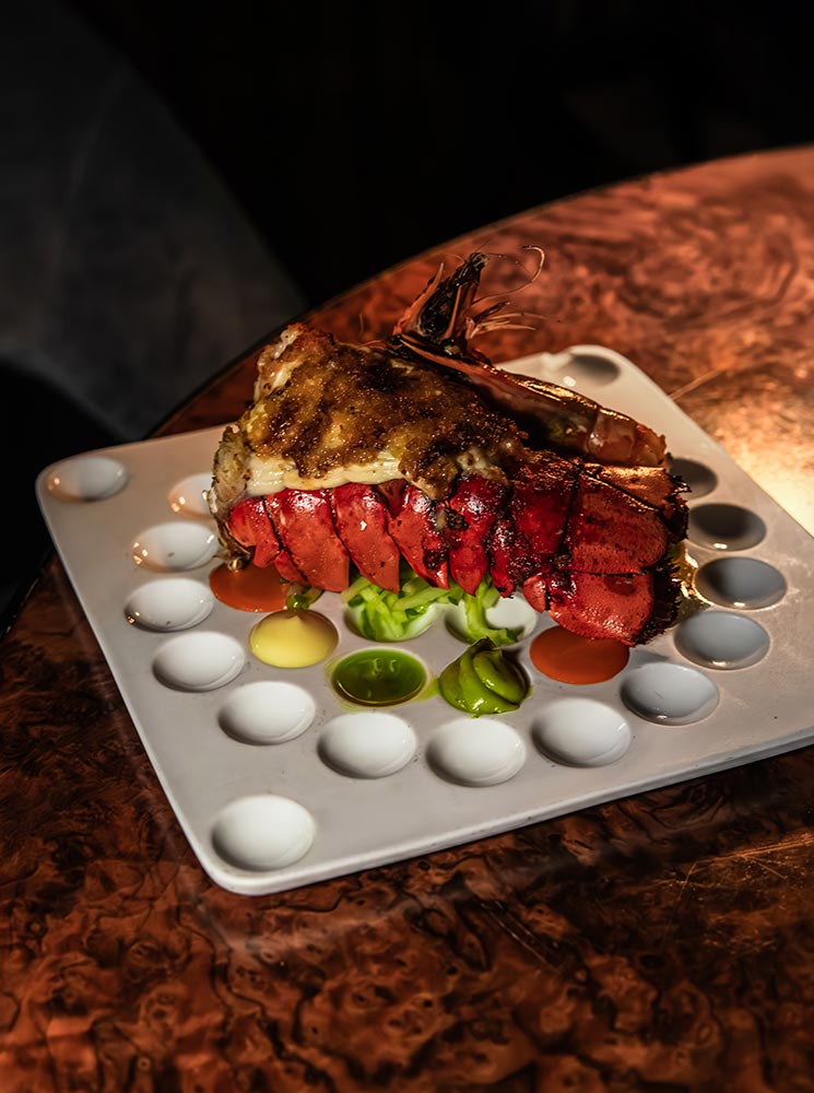 This image showcases a beautifully prepared lobster tail served at Apotek in Reykjavik, presented on a unique plate with various dipping sauces in individual compartments. The lobster is grilled to perfection with a golden glaze, accompanied by fresh greens. The warm lighting accentuates the dish's luxurious appeal.