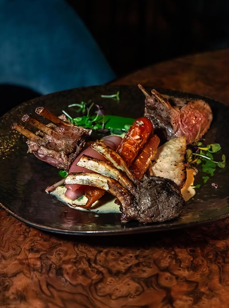 This image features a succulent rack of lamb served at Apotek in Reykjavik, beautifully plated with perfectly grilled chops, roasted vegetables, and a creamy sauce drizzled for garnish. The rich textures and warm tones of the dish highlight its gourmet presentation, creating a luxurious and inviting dining experience.