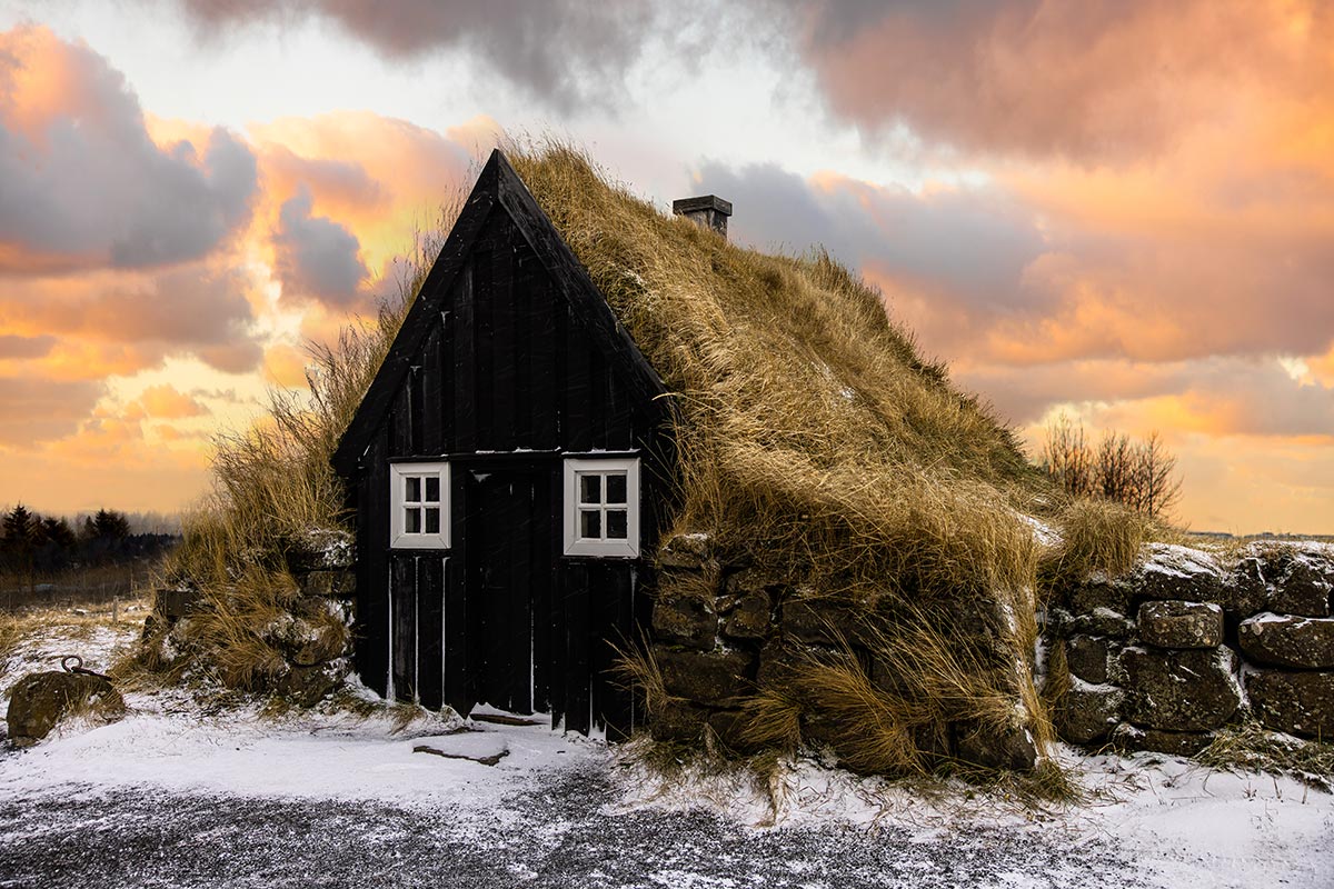 This image features a traditional Icelandic turf house at Árbær Open Air Museum in Reykjavik. The black wooden structure is covered with a thick, grassy roof, blending harmoniously into the winter landscape under a vibrant sunset. It showcases a glimpse into Iceland's historic architectural heritage.