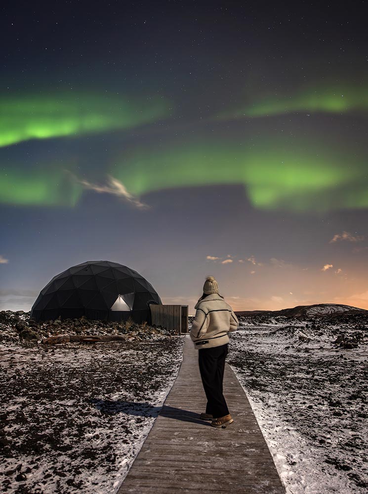 This image captures a person walking on a wooden path toward a geodesic dome under a vibrant display of the Northern Lights at the Aurora Basecamp near Reykjavik. The snow-dusted ground and the glowing green aurora overhead create a magical, otherworldly atmosphere, blending natural beauty with the serene structure in the foreground.