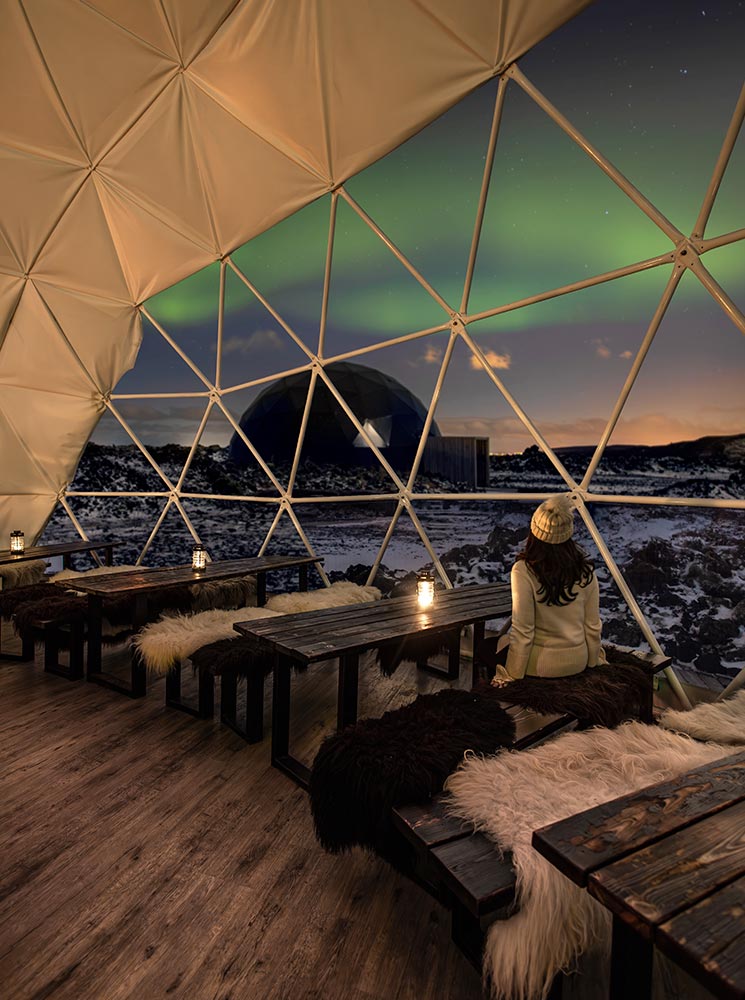 This image captures the cozy interior of a geodesic dome at Aurora Basecamp near Reykjavik, with fur-lined wooden benches and lanterns creating a warm, inviting ambiance. Through the dome’s triangular glass panels, the vibrant green Northern Lights and a snow-covered landscape are visible, blending comfort with the beauty of Iceland's natural wonders.