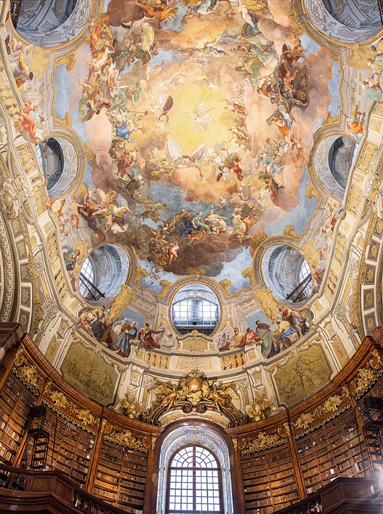 This image highlights the ornate ceiling fresco in the State Hall of the Austrian National Library, Vienna, depicting vibrant mythological and celestial scenes radiating from a central burst of light. The grand circular mural is surrounded by arched windows and framed by gilded architectural details, with towering bookshelves beneath. The artwork and intricate details create a sense of awe, embodying the cultural and artistic heritage of this historic library.