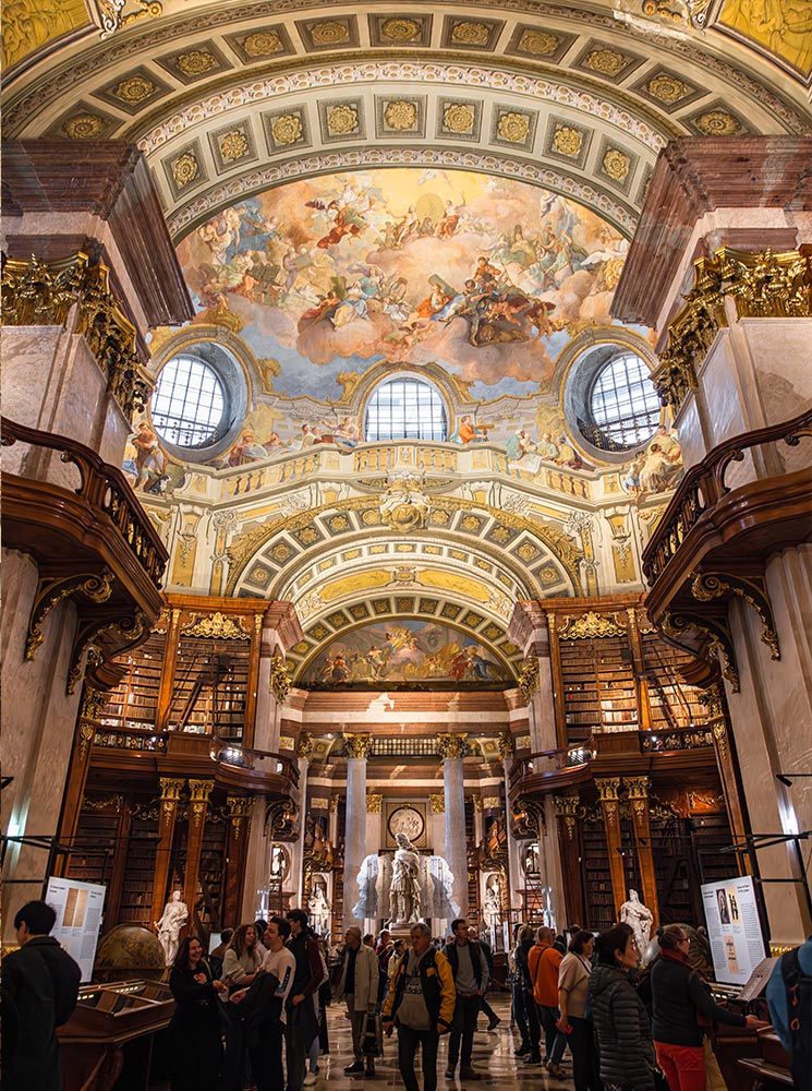 This image captures the magnificent State Hall of the Austrian National Library in Vienna, with its grandiose Baroque architecture, towering bookshelves, and intricate ceiling fresco depicting mythological scenes. The hall is illuminated by natural light through large arched windows and features statues, marble pillars, and ornate gold detailing. Visitors admire the opulent space, which epitomizes the grandeur of historical European libraries.