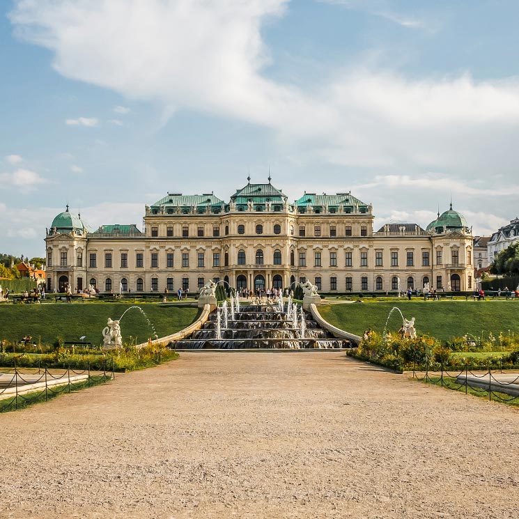 This image presents Vienna's Belvedere Palace, a grand baroque structure with green copper domes, standing proudly under a bright sky. In the foreground, a tiered fountain with sculpted figures cascades down towards the viewer, surrounded by manicured gardens that add to the palace's majestic allure. Visitors stroll through the grounds, highlighting the palace as both a historical monument and a scenic destination.