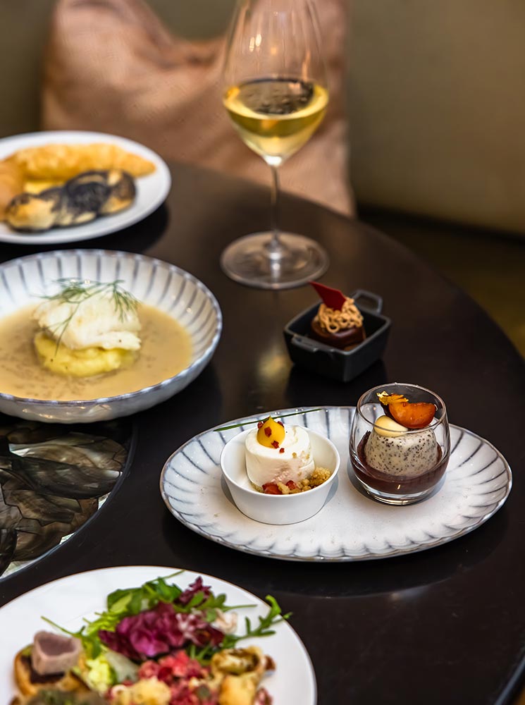 This image showcases a refined brunch spread at The Bank Brasserie & Bar, featuring an assortment of elegantly plated dishes. Delicate desserts, a fresh salad, a warm soup, and a glass of white wine are artfully arranged on a dark table. The presentation highlights the sophistication and culinary finesse of the brunch experience at this upscale Vienna restaurant.