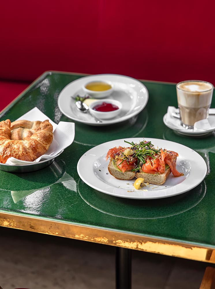 This image captures a stylish brunch setting at Brasserie Palmenhaus in Vienna, featuring a plate of smoked salmon on toast topped with microgreens, a side of croissants, and a latte. A dish of jam and butter completes the meal, all served on a glossy green table with gold trim against a red banquette backdrop. The spread exudes a refined, European café atmosphere perfect for a leisurely brunch.