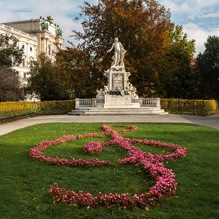 This image showcases the Mozart Monument in Vienna's Burggarten, featuring a statue of the composer atop a pedestal adorned with relief sculptures. In front of the monument, a beautifully arranged flower bed in the shape of a treble clef, made of vibrant pink blooms, stands out against the green grass, celebrating Mozart's legacy in a picturesque garden setting surrounded by autumn-colored trees.