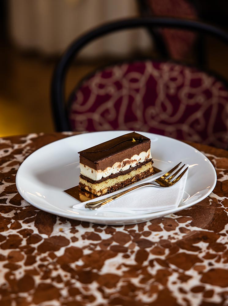 This image features a slice of layered chocolate cake from Café Central in Vienna, artfully plated on a white dish with a fork and napkin. The cake has multiple layers, including creamy and chocolate elements, topped with a glossy chocolate glaze. The patterned tabletop and plush chair in the background add a touch of elegance, highlighting the luxurious Viennese café experience.