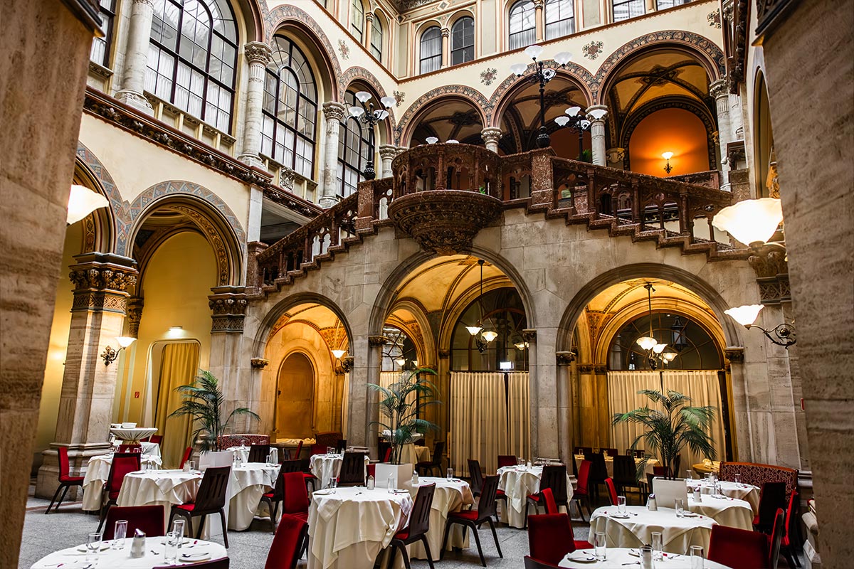 This image displays the elegant interior of Café Central in Vienna, with round tables covered in white tablecloths and set with red chairs. The high, arched ceilings, ornate staircase, and vintage lamps contribute to the café's historic and refined ambiance. The combination of warm lighting and architectural details creates a classic Viennese charm, making it an inviting space for patrons.






