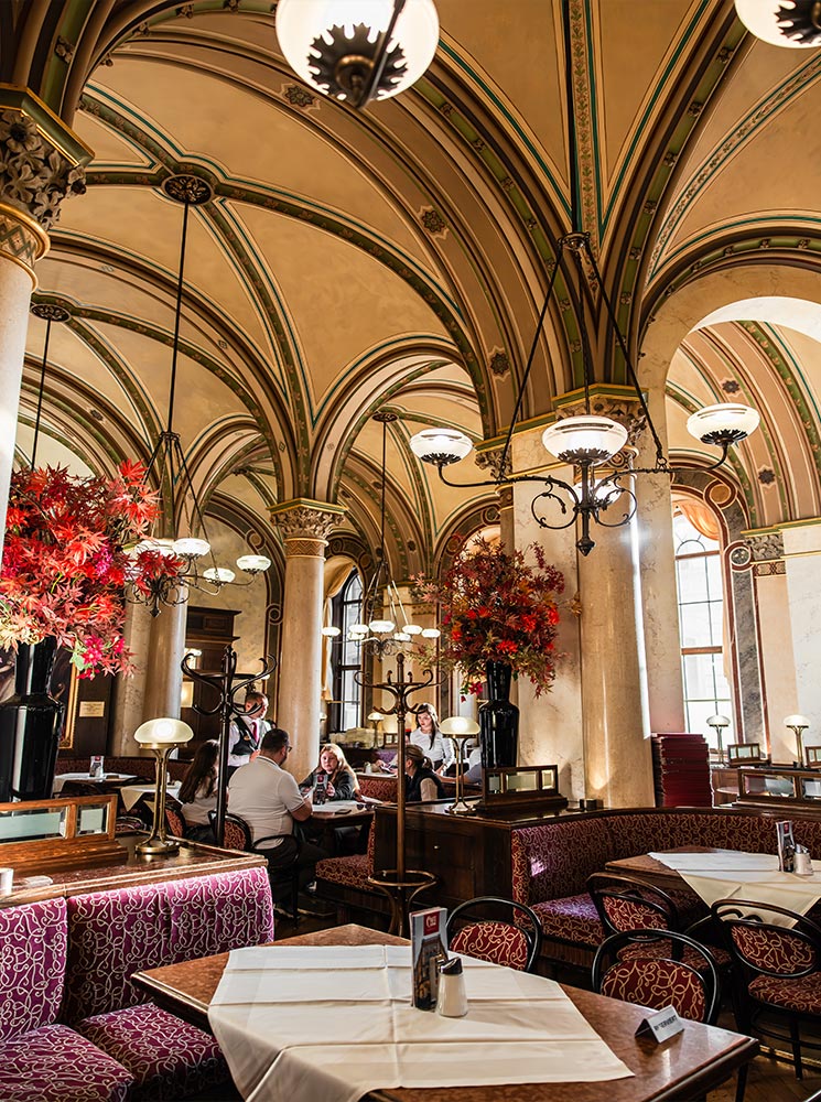 This image shows the interior of Café Central in Vienna, highlighting its vaulted ceilings with intricate green and gold detailing, and warm, ornate lighting. Red floral arrangements and plush seating with patterned upholstery create a cozy yet elegant atmosphere. Patrons are seen seated at tables, enjoying the café's inviting and historic ambiance.