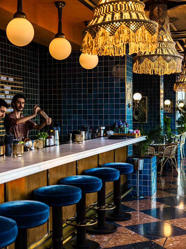 This image shows the interior bar area of Cayo Coco Rooftop Bar in Vienna, featuring deep blue tiled walls, a sleek marble countertop, and plush velvet barstools. Ornate hanging lamps with fringe details and globe lights create a warm, ambient glow, enhancing the bar's tropical and luxurious aesthetic. A bartender is seen crafting a drink, adding to the lively and inviting atmosphere of this stylish venue.