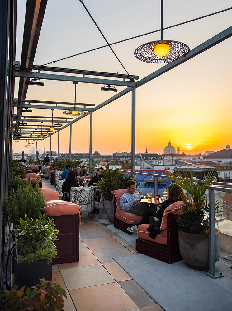 This image captures the vibrant sunset view from Cayo Coco Rooftop Bar, where patrons relax on plush seating surrounded by greenery. The terrace is adorned with hanging woven lamps that cast a warm glow as the sun sets over the Vienna skyline. The mix of comfortable seating, modern decor, and stunning views creates an inviting ambiance for evening socializing.