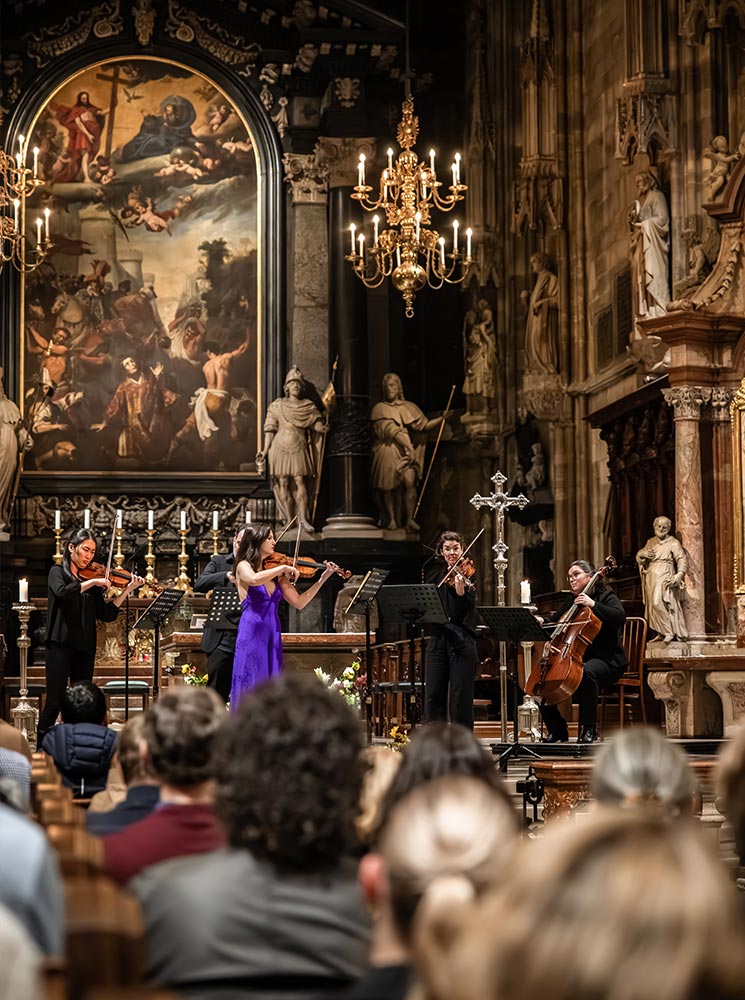 This image captures a classical concert inside St. Stephen's Cathedral in Vienna, featuring a quartet of musicians performing before an attentive audience. The ornate altar backdrop includes a large religious painting, marble statues, and golden chandeliers that cast a warm glow over the scene. The ambiance combines the reverence of the historic cathedral with the elegance of live classical music.