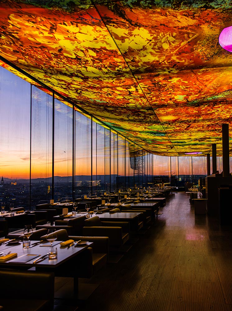 This image showcases the vibrant rooftop dining area of Das LOFT in Vienna, featuring floor-to-ceiling windows with panoramic city views at sunset. The ceiling is adorned with colorful, abstract artwork that casts a warm glow over the modern, neatly set tables below. The combination of artistic ambiance and breathtaking views creates a sophisticated and memorable dining experience.