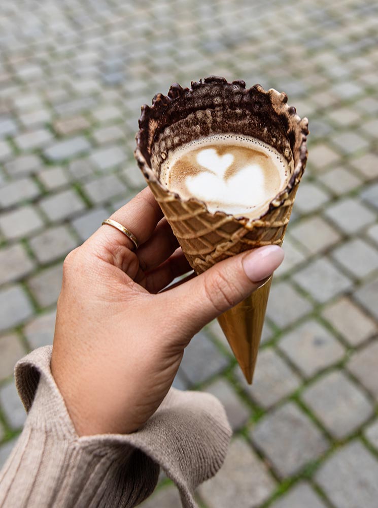 This image features a unique coffee presentation from Fenster Café in Vienna, served in a chocolate-lined waffle cone with a heart-shaped latte art on top. Held by a hand with a cozy sweater sleeve, the coffee cone stands out against the cobblestone background, blending a creative twist on coffee with an urban European setting.