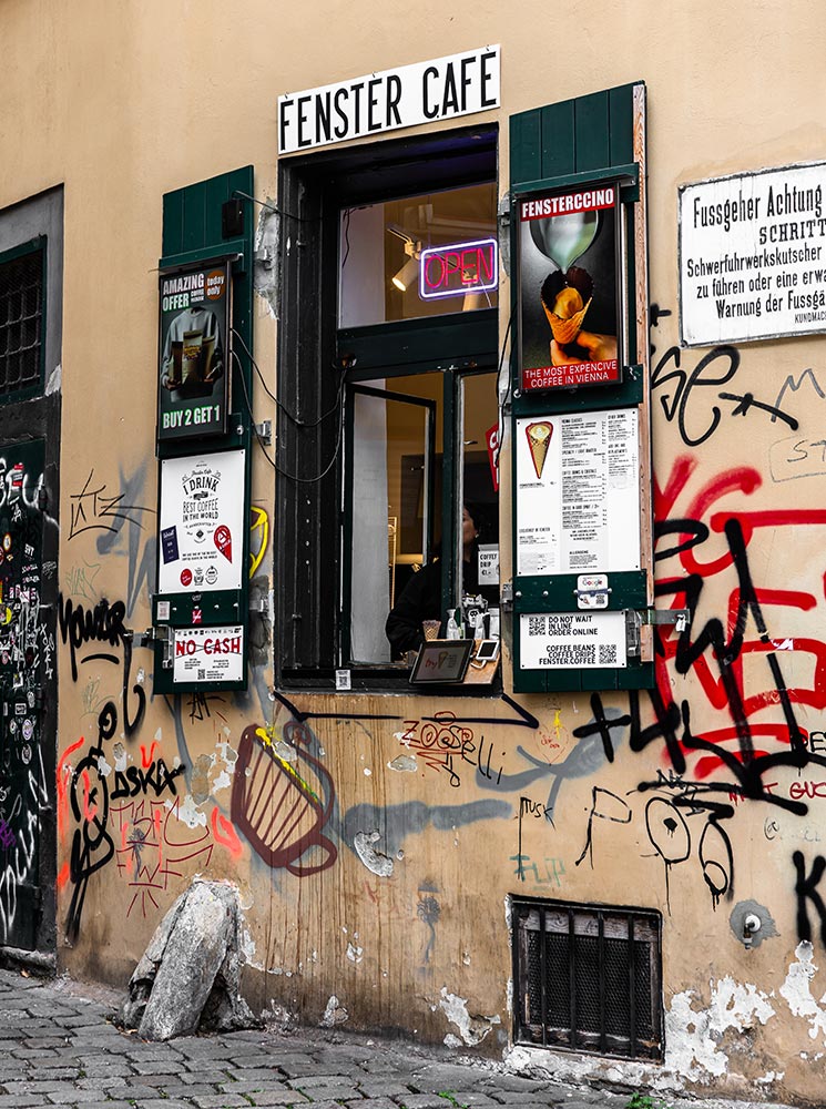 This image depicts the exterior of Fenster Café in Vienna, a small coffee spot known for its "coffee in a cone" served through a window. The storefront is surrounded by colorful graffiti, signage advertising the café's unique offerings, and a neon "OPEN" sign, creating a blend of urban grit and local charm. The eclectic decor highlights the café's unconventional appeal as a hidden gem in the city.