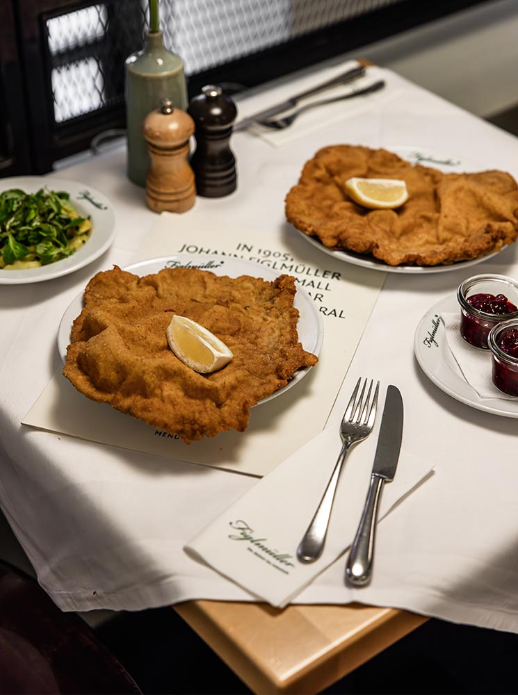 This image presents a classic Viennese meal at Figlmüller, featuring two large Wiener Schnitzels, each garnished with a lemon wedge. The table is set with a side of salad, a dish of cranberry sauce, and traditional cutlery on a branded napkin, all atop a pristine white tablecloth. The scene captures the iconic Viennese dining experience with a focus on authentic presentation and generous portions.