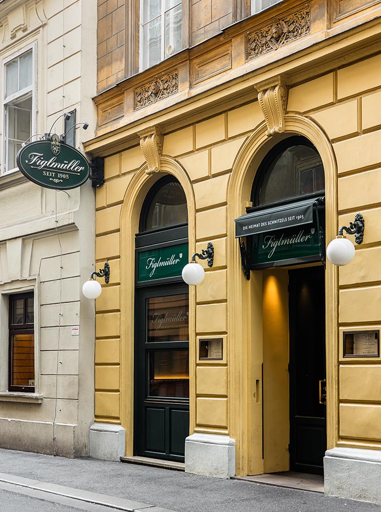 This image shows the exterior of Figlmüller, a renowned restaurant in Vienna famous for its Wiener Schnitzel, established in 1905. The building features a classic yellow facade with green signage above arched doorways, and round white lights on either side, giving it a traditional and inviting appearance. The atmosphere reflects the restaurant’s historical roots and prominence in Viennese cuisine.