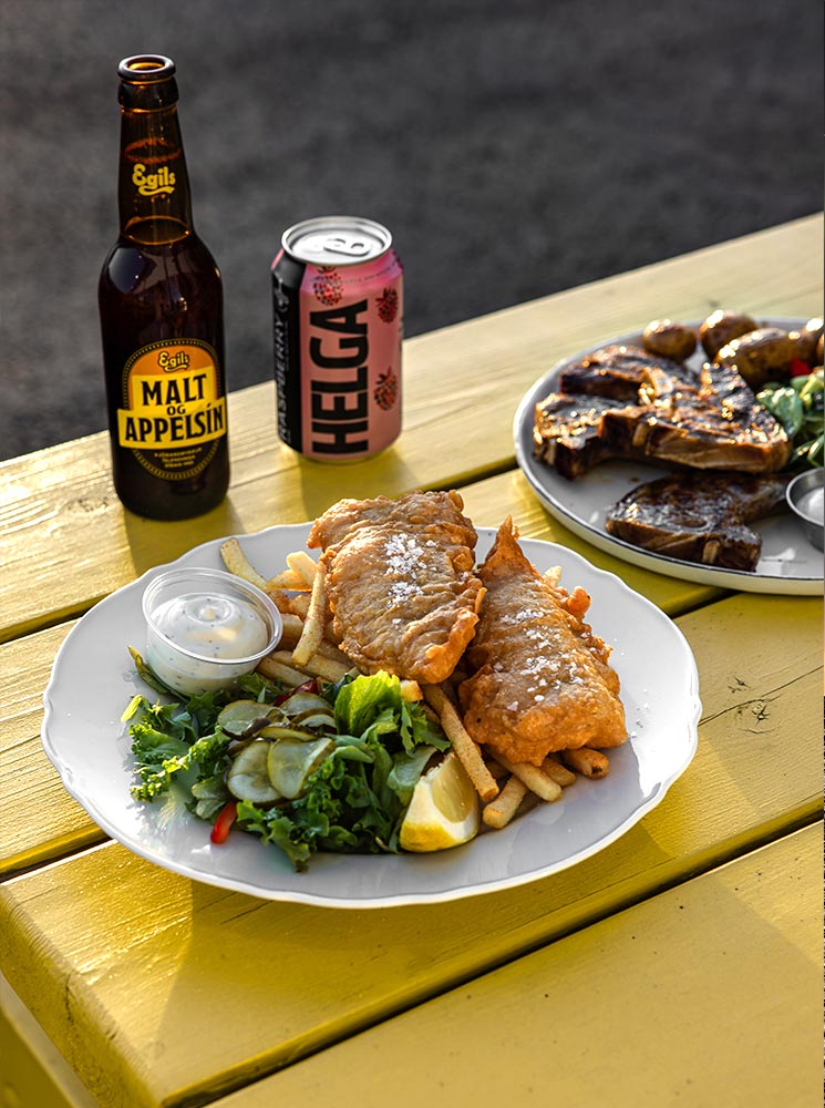 This image showcases a delicious serving of fish and chips from Grandi Food Hall in Reykjavik, featuring golden-battered fish, crispy fries, a fresh side salad, and a slice of lemon, accompanied by tartar sauce. The meal is set on a sunny yellow picnic table, with beverages including Egils Malt og Appelsín and a can of Helga soda adding a local Icelandic touch to the casual dining experience.