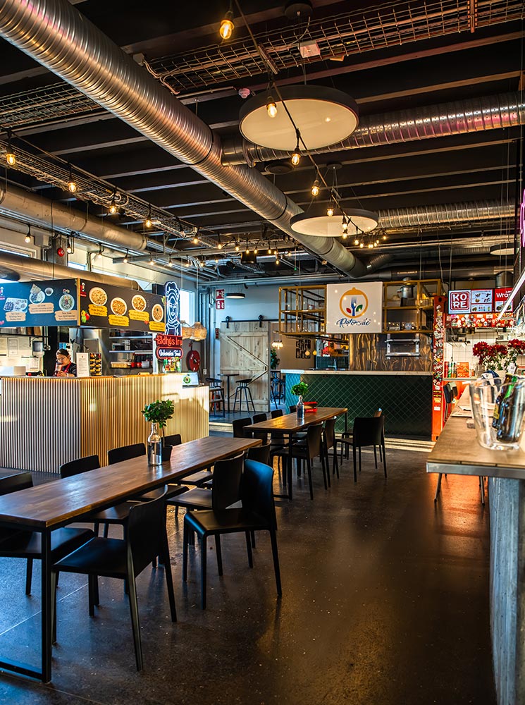 This image captures the inviting interior of the Grandi Food Hall in Reykjavik, featuring a variety of food stalls and a modern industrial design. Wooden communal tables with simple floral centerpieces are complemented by exposed ceilings with ductwork and hanging lights, creating a trendy yet relaxed atmosphere. The vibrant signage and diverse dining options hint at a lively and eclectic culinary experience.