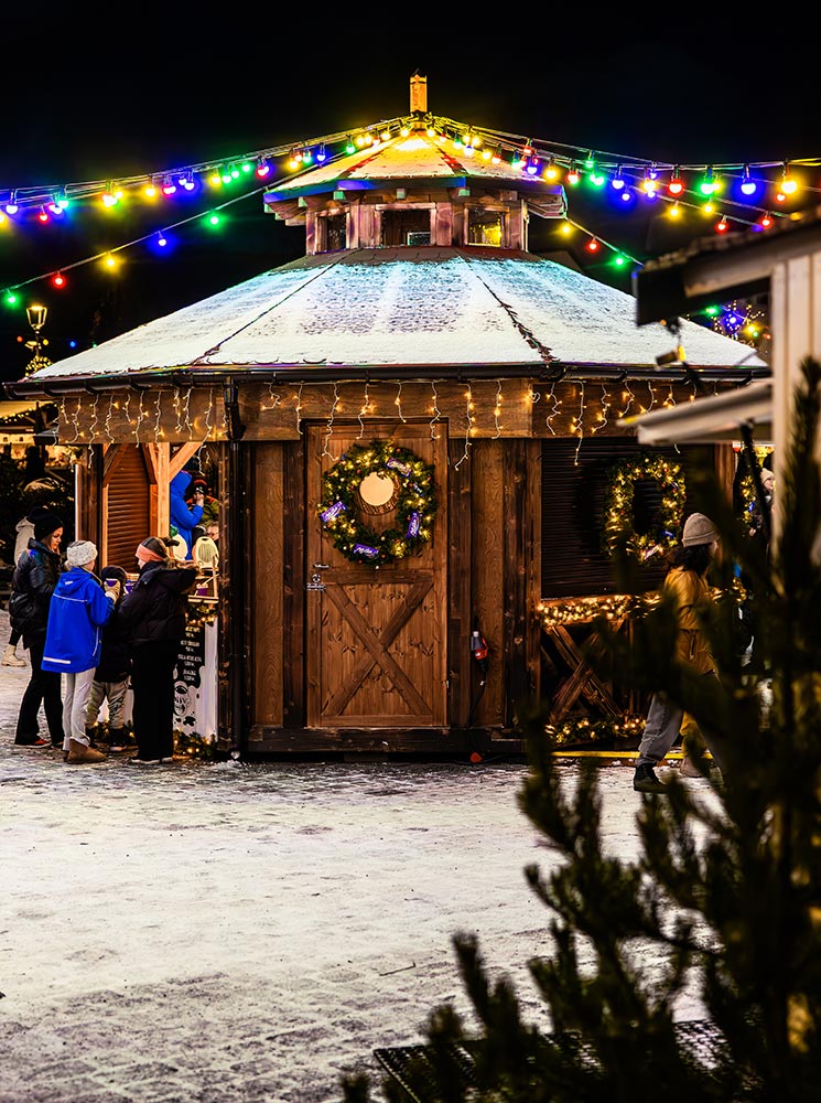 This image captures a festive Christmas market scene in Hafnarfjörður near Reykjavik, featuring a wooden stall adorned with wreaths, string lights, and a snowy roof. Vibrant multicolored lights are strung overhead, creating a cheerful ambiance as people gather around to enjoy the holiday atmosphere. The cozy setting is filled with warmth and holiday spirit despite the chilly weather.