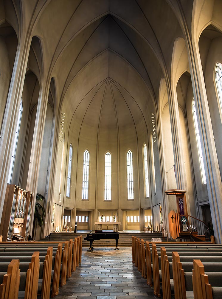 This image captures the serene interior of Hallgrímskirkja in Reykjavik, showcasing its soaring, minimalist Gothic arches and rows of wooden pews. A grand piano is positioned near the altar, surrounded by soft natural light streaming through tall, narrow windows, emphasizing the church's elegant simplicity and spiritual ambiance.