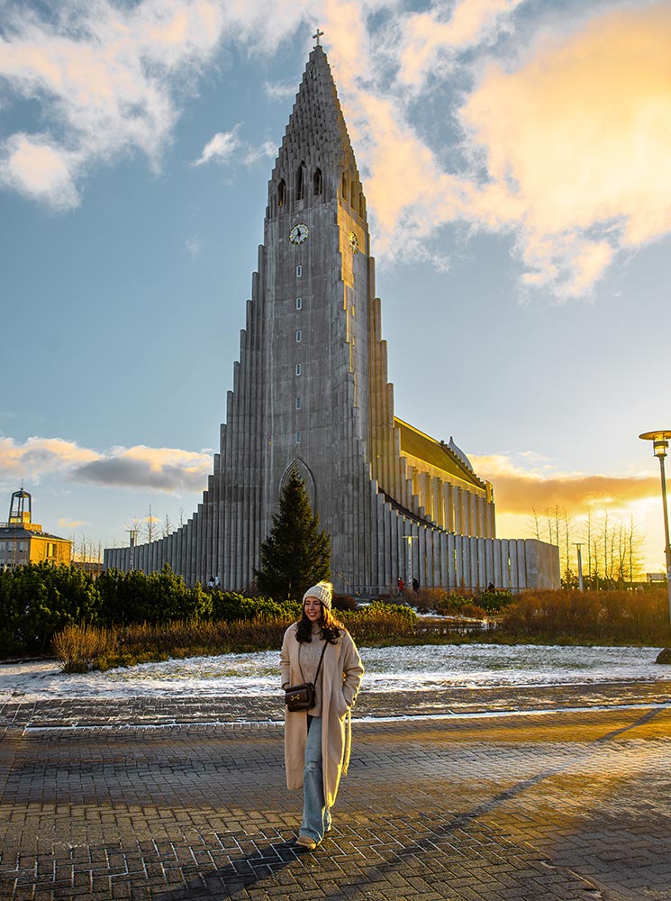 This image features Hallgrímskirkja, the iconic church in Reykjavik, bathed in golden sunlight under a partly cloudy sky. A smiling woman dressed in a cozy coat and knit hat walks in the foreground on a lightly snow-dusted path, adding warmth and life to the scene.