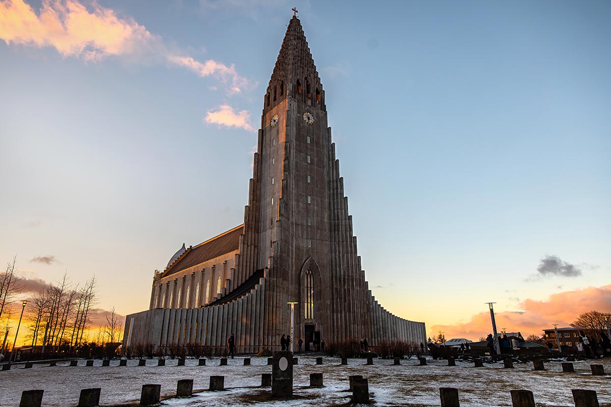 This image depicts Hallgrímskirkja, a striking concrete church in Reykjavik, Iceland, with its iconic stepped facade and towering spire, captured during a serene sunset. The foreground shows a snow-dusted plaza with scattered stone pillars, enhancing the architectural grandeur against the colorful sky.