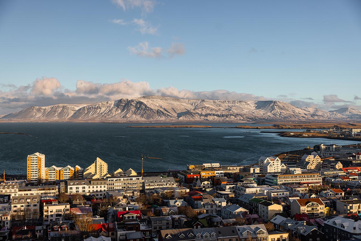 This image captures a panoramic view of Reykjavik from the tower of Hallgrímskirkja, showcasing colorful rooftops, modern architecture, and the surrounding icy waters. In the background, the snow-capped Esja mountain range rises dramatically under a clear blue sky, completing this breathtaking Icelandic landscape.