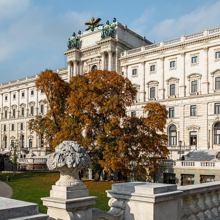 This image displays the historic Hofburg Palace in Vienna, set against a backdrop of blue skies and autumn foliage. The palace's grand architecture is accentuated by classical statues, ornate columns, and a rooftop adorned with statues of chariots. The warm colors of the trees add a seasonal charm to the imperial ambiance of this iconic landmark.