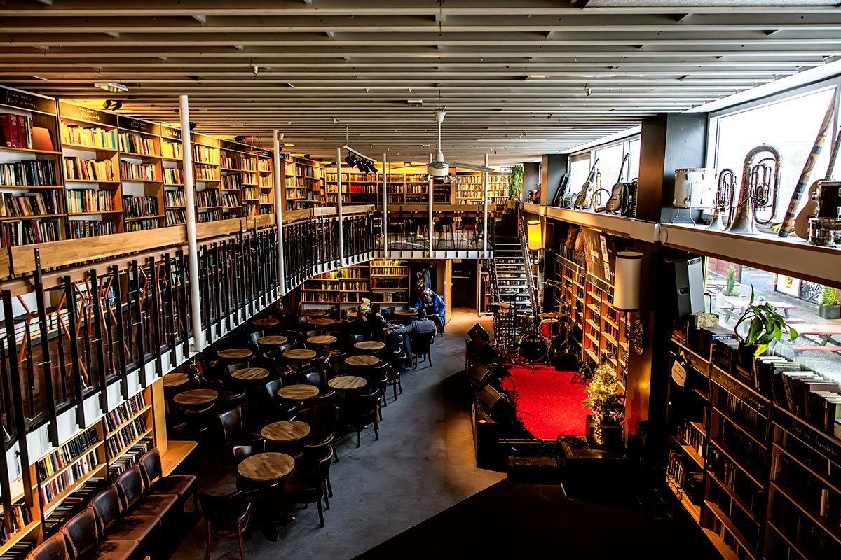 This image showcases the interior of Hús Máls og Menningar, a unique Reykjavik venue that blends a bookstore with a cozy café and a live performance space. Shelves filled with books span two levels, encircling a central seating area and a small stage with musical instruments ready for performances. The warm, inviting ambiance highlights its role as a cultural and social hub.