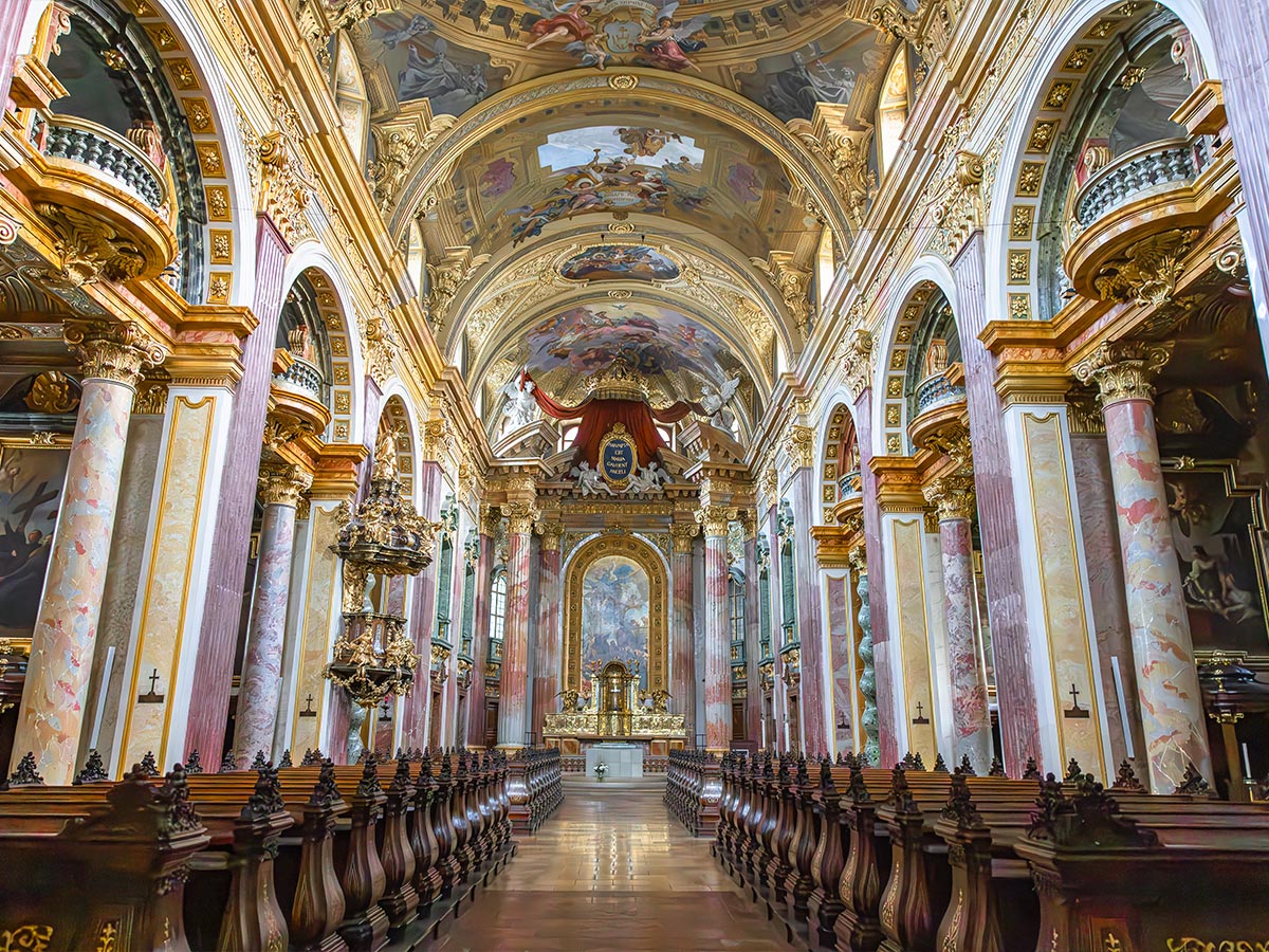 This image captures the breathtaking interior of the Jesuit Church in Vienna, adorned with towering marble columns, intricate gold accents, and richly painted frescoes on the arched ceiling. The aisle is flanked by dark wooden pews leading toward an ornate altar, framed by lavish decorations that reflect the Baroque opulence of the church. The grand design and elaborate details create a sense of awe and reverence in this historic space.