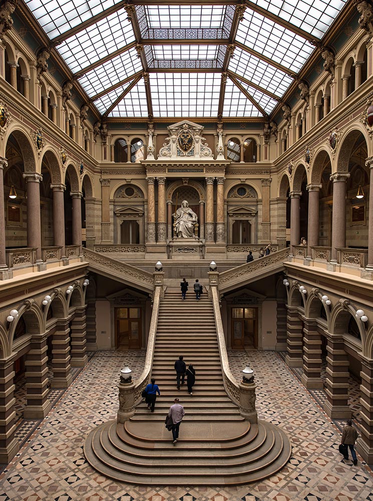 This image captures the grand interior of the Palace of Justice in Vienna, with its impressive central staircase leading up to a statue set within an ornate archway. The hall is surrounded by arched balconies and columns, all under a glass ceiling that fills the space with natural light. The intricate tile floor and classical architectural details emphasize the building's historical and majestic atmosphere.