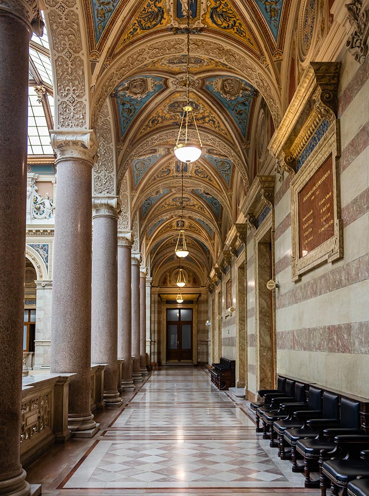 This image features a beautifully detailed corridor inside Vienna's Palace of Justice, with tall marble columns, arched ceilings adorned with intricate blue and gold frescoes, and elegant hanging lights. The polished marble floor reflects the refined architecture, while a row of classic black chairs lines one wall, adding to the stately ambiance of this historic building. The corridor’s design exemplifies European grandeur and legal tradition.