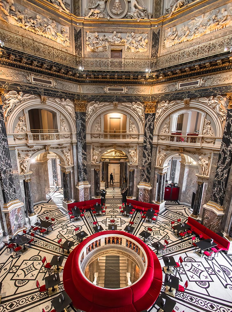 This image shows the elegant interior of the Kunsthistorisches Museum café in Vienna, with its marble columns, intricate wall carvings, and grand architectural details. The café seating area, featuring circular red velvet benches and black tables, is set against a beautiful mosaic floor, creating a refined and artistic atmosphere. The high ceiling is adorned with classical reliefs and gilded accents, adding to the museum's grandeur.