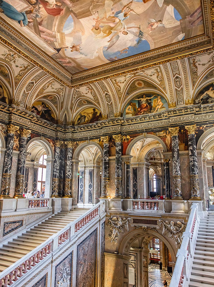 This image shows the elegant interior of the Kunsthistorisches Museum café in Vienna, with its marble columns, intricate wall carvings, and grand architectural details. The café seating area, featuring circular red velvet benches and black tables, is set against a beautiful mosaic floor, creating a refined and artistic atmosphere. The high ceiling is adorned with classical reliefs and gilded accents, adding to the museum's grandeur.