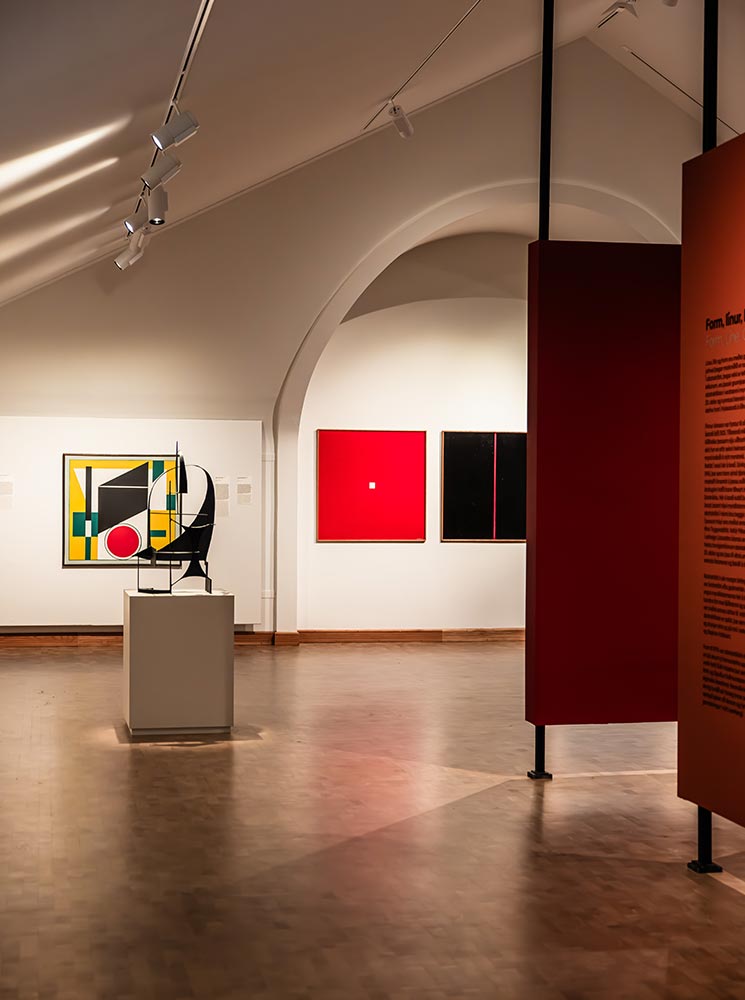 This image showcases a modern exhibition at the National Gallery of Iceland, featuring bold, abstract artworks. The space includes geometric paintings with striking color contrasts, such as red, black, and yellow, alongside a minimalist sculpture on a pedestal. Clean lines, arching ceilings, and focused lighting enhance the contemporary and thought-provoking atmosphere.