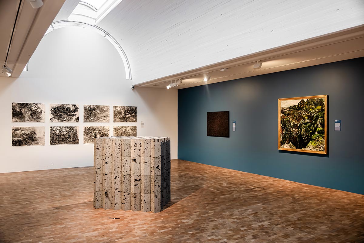 This image shows an exhibition space in the National Gallery of Iceland, featuring a mix of modern and traditional artworks. The gallery includes a centerpiece sculpture made of textured stone blocks, a series of monochrome drawings on a white wall, and a colorful framed landscape painting displayed on a blue accent wall. The bright lighting and high ceiling create a serene and spacious atmosphere.