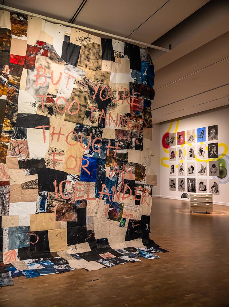 This image captures an installation at the National Gallery of Iceland, featuring a large textile artwork with the text "BUT YOU'RE TOO TANNED THOUGH FOR AN ICELANDER" written across it. The piece is a patchwork of painted and dyed fabric, displayed alongside a series of black-and-white portraits and abstract wall art in the background. The exhibit evokes a mix of cultural commentary and artistic exploration within a modern gallery setting.