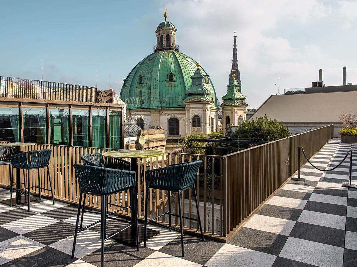 This image features the rooftop terrace of Neue Hoheit Brasserie in Vienna, with modern seating and a checkered tile floor that overlooks the city. The view includes the iconic green dome of a nearby historic building, creating a contrast between contemporary design and classic Viennese architecture. The open-air setting offers a scenic, elevated spot to enjoy the cityscape under a clear sky.