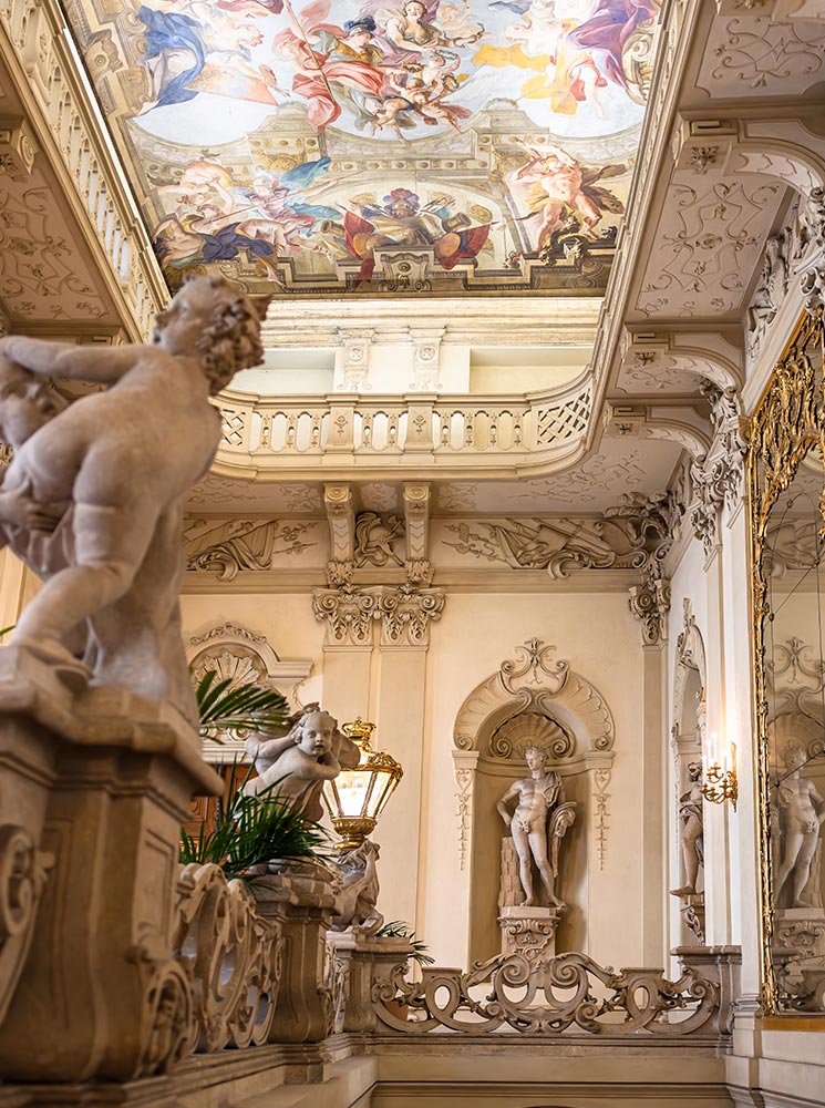 This image captures a detailed view of the ornate interior of Palais Daun-Kinsky, focusing on a marble statue, decorative molding, and an elaborate ceiling fresco. The fresco features vivid mythological scenes, adding to the elegance of the space, while stone cherubs and a grand lamp lend a touch of classical artistry. The intricate architecture and rich decor embody the palace's luxurious, historic charm.