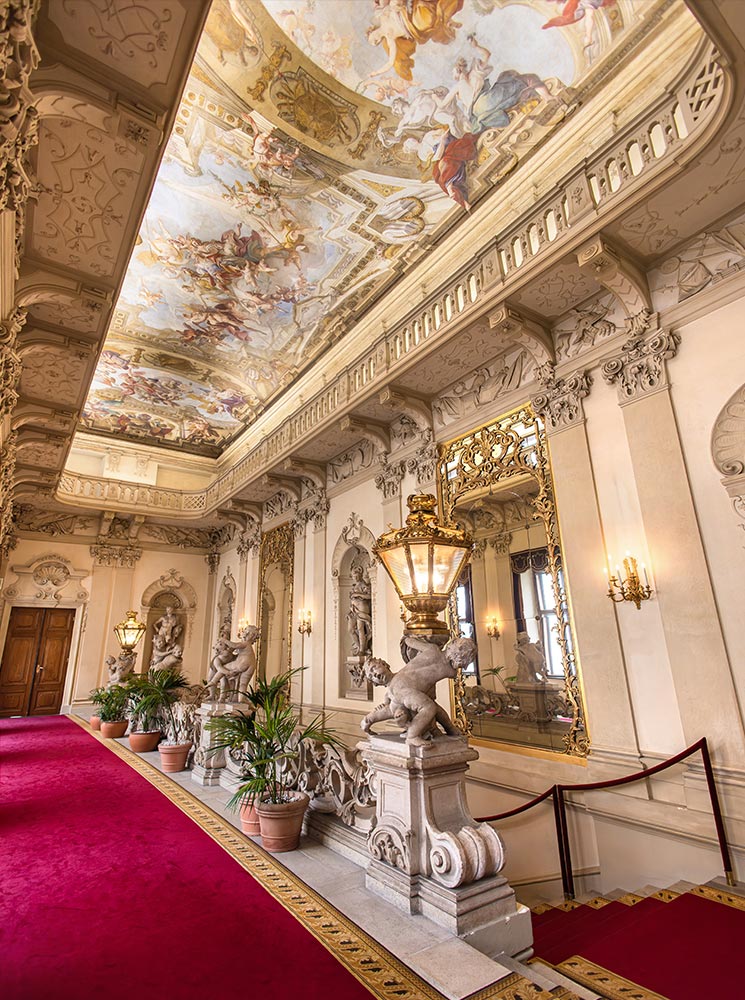 This image captures the luxurious interior of Palais Daun-Kinsky in Vienna, with its red carpeted corridor, ornate statues, and lavish ceiling frescoes depicting mythological scenes. The walls are adorned with intricate moldings, gilded mirrors, and classic light fixtures, enhancing the opulent ambiance. The decorative stone figures and lush potted plants add to the regal aesthetic of this historic palace.