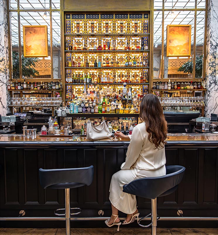 This image captures an elegant bar scene at the Park Hyatt Vienna, where a woman in a chic white outfit sits at the bar, holding a glass. The bar boasts an impressive backlit display of spirits and intricate stained glass detailing, creating a warm, luxurious ambiance ideal for a sophisticated evening out.